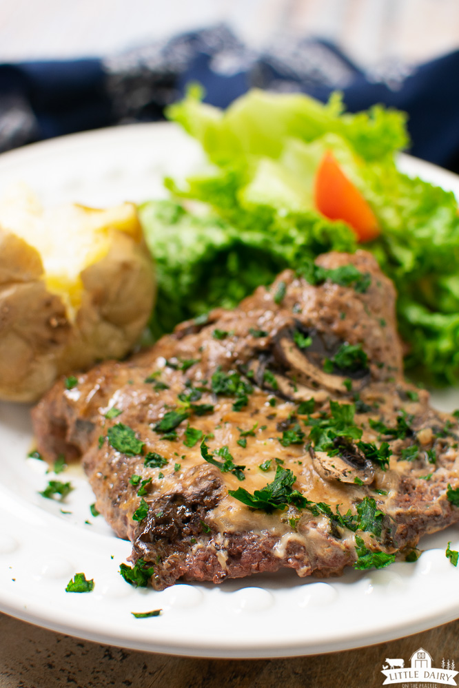 a white plate with green salad, a baked potato, and cooked cubed steaks with a mushroom gravy on top