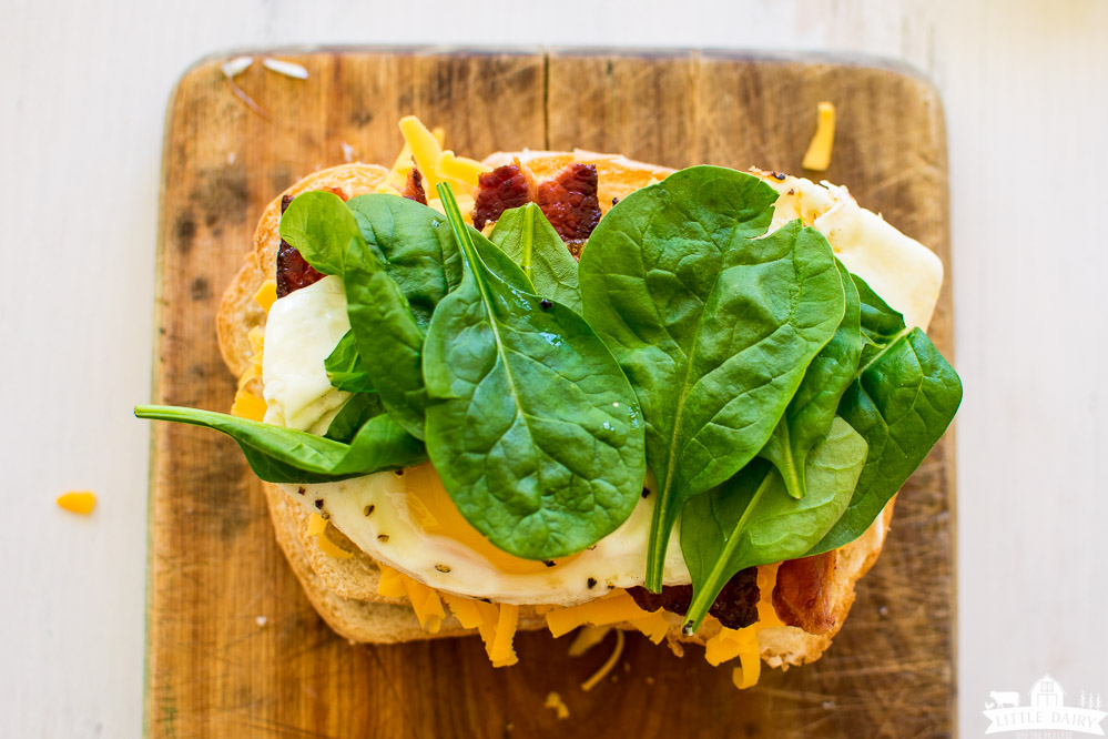 a piece of bread stacked with grated cheese, a fried egg, bacon, and baby spinach