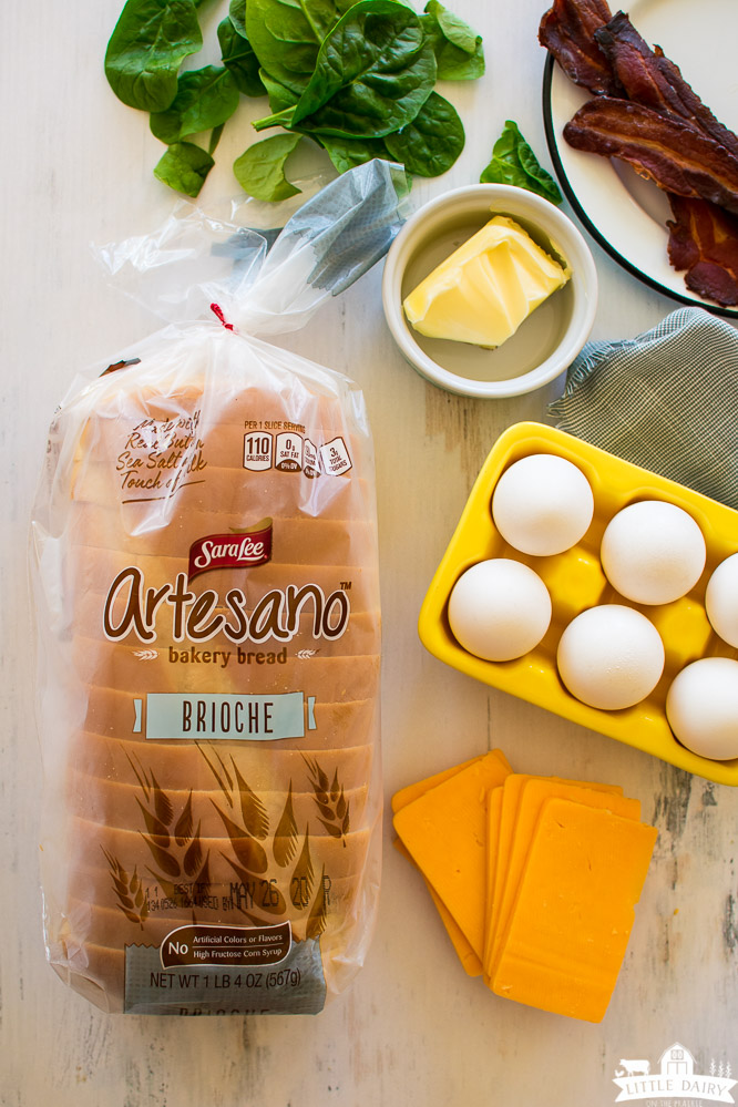 ingredients need to make fried egg sandwich- Sara Lee Artesano bread in the package, eggs in a yellow egg crate, butter in a dish, fried bacon on a plate, fresh spinach, and sliced cheddar cheese