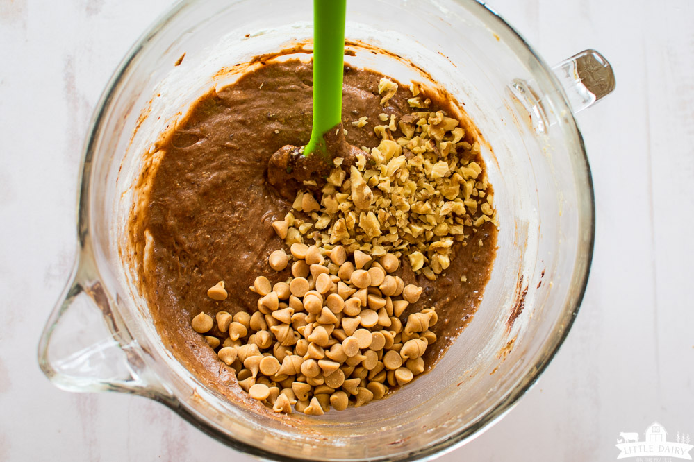 brownie batter in a glass mixing bowl topped with peanut butter chips and chopped walnuts with a green spatula
