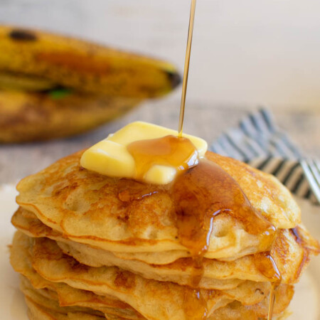 a stack of pancakes with a pat of butter on top, maple syrup being drizzled over the top