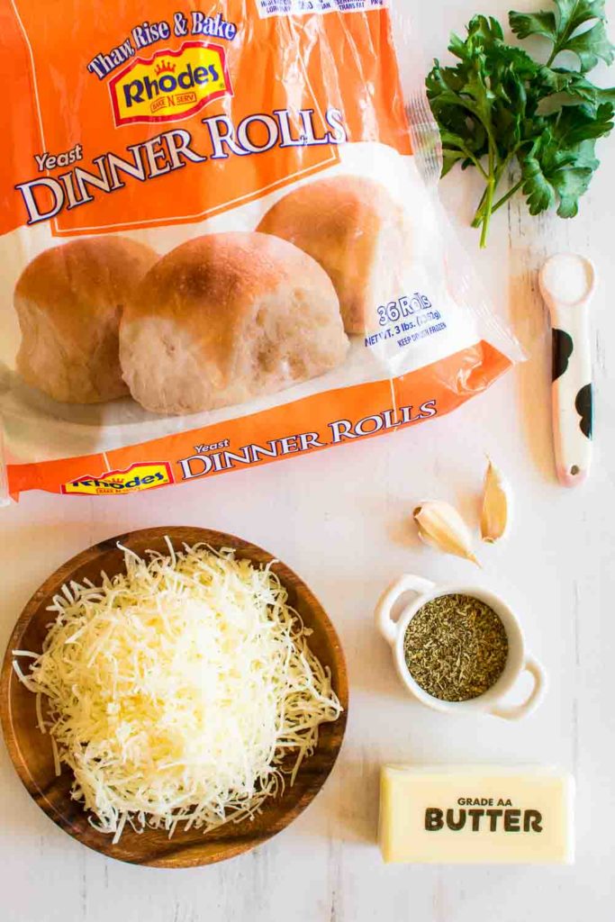 ingredients needed to make the recipe; grated parmesan in a wooden bowl, fresh parsley, a bag of frozen dinner rolls, Italian seasoning, salt, garlic, and butter