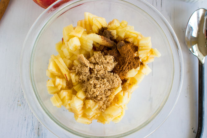 peeled and diced apples in a glass bowl topped with cinnamon, brown sugar, and spices