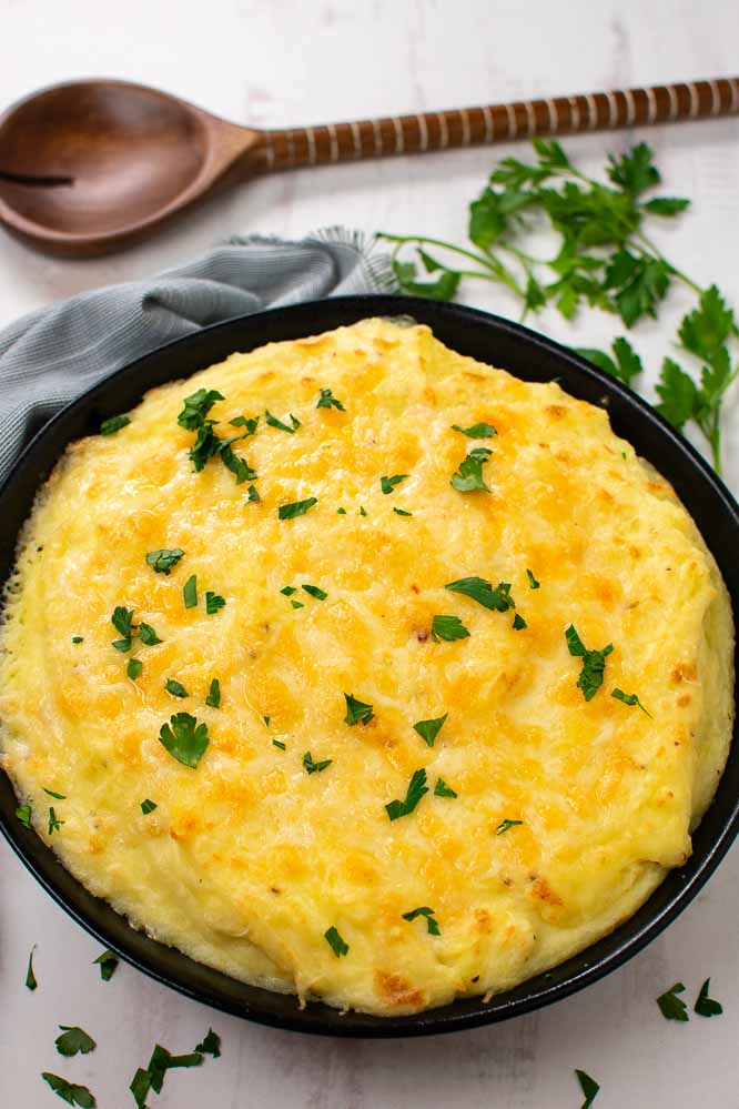 a black bowl with mashed potatoes and a layer of melted cheese on top with a wooden spoon and fresh parsley