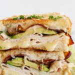 a hot turkey sandwich cut in half and topped with avocado, turkey, and bacon