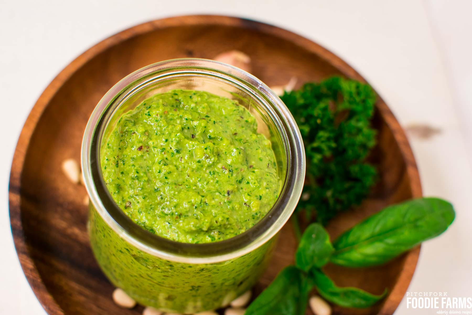 A jar of green pesto with fresh basil leaves, pine nuts and parsley sprinkled around it on a wooden plate.