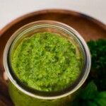 A glass jar with green pesto on a wooden plate.