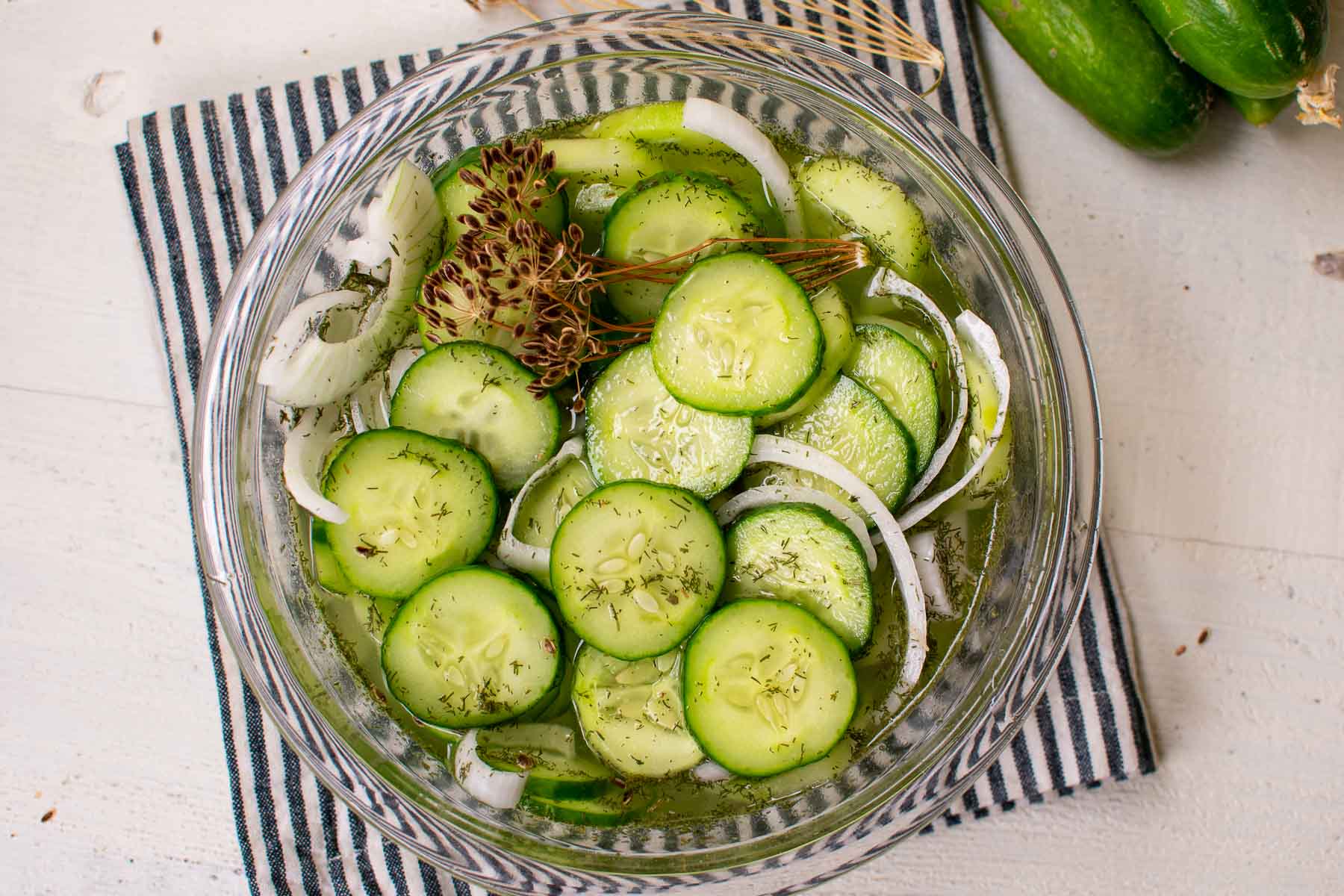 Sliced cucumbers in a glass bowl with veggies on a striped napkin.