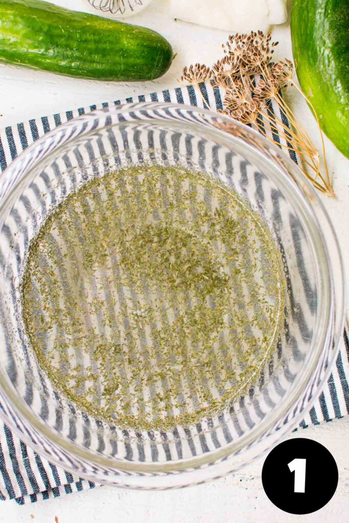 Marinade with fresh herbs in a glass bowl with cucumbers and dill weed in the back.