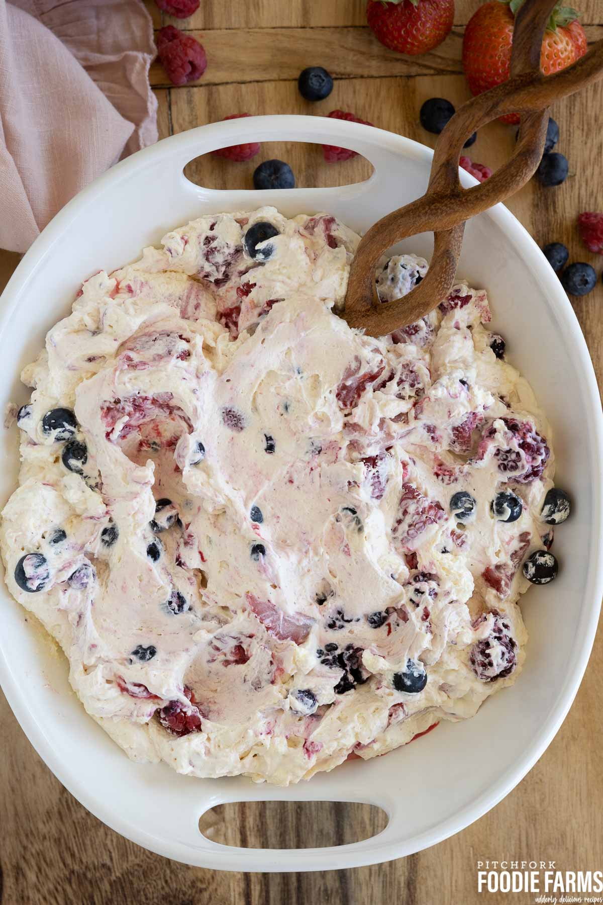 Birdseye view of a creamy fruit salad with raspberries, blueberries, and strawberries in a white bowl with a wooden spoon.