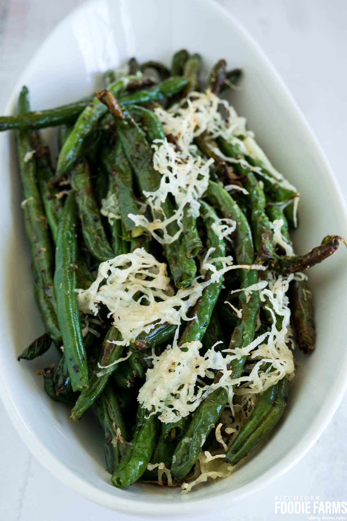 Oven baked green beans in a white serving dish with toasted parmesan cheese on top.