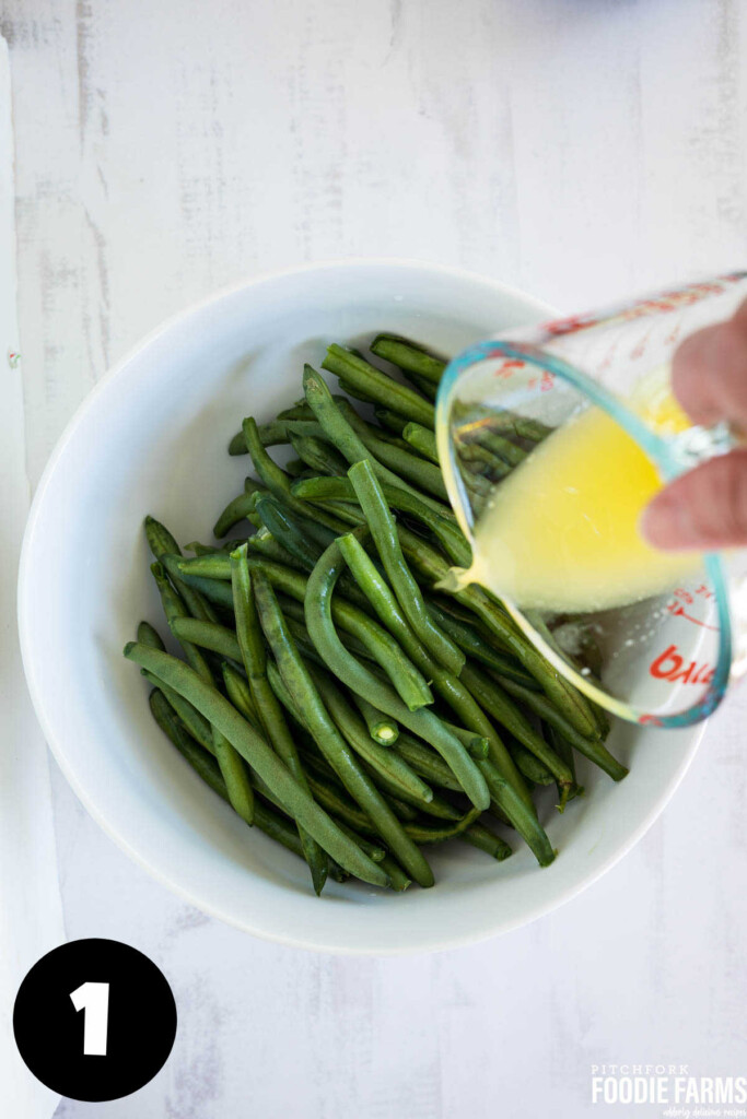 A white bowl with green beans and a measuring cup of melted butter being drizzled in.