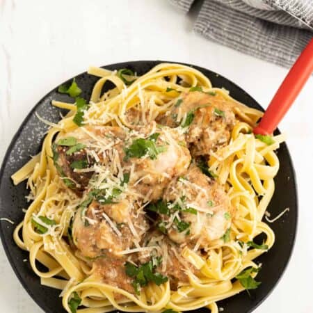 Pasta topped with meatballs in a creamy Italian tomato sauce sprinkled with cheese and herbs on a black plate.