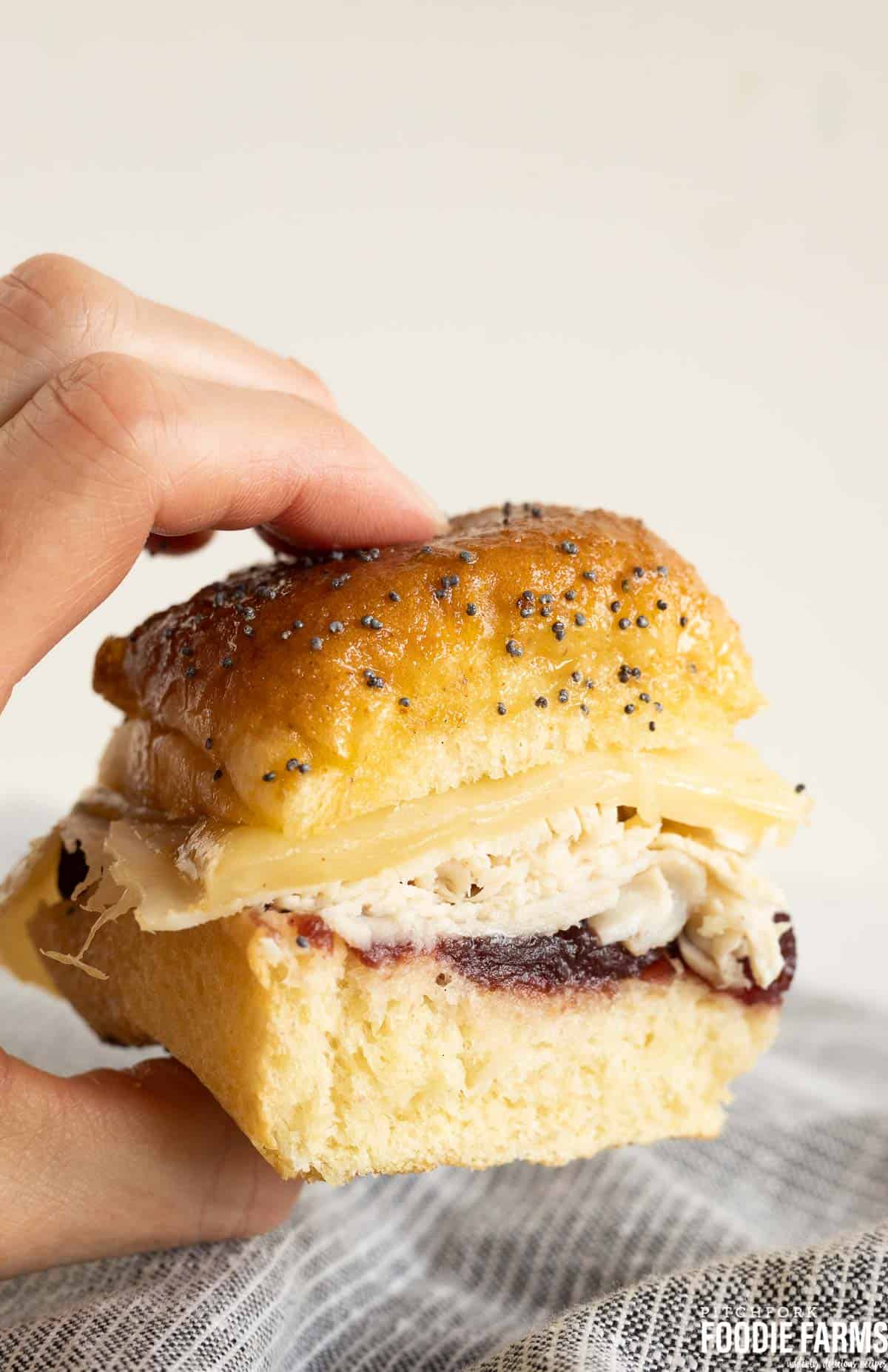A hand holding a baked slider on a Hawaiian roll with deli turkey, cheese, and cranberry sauce.