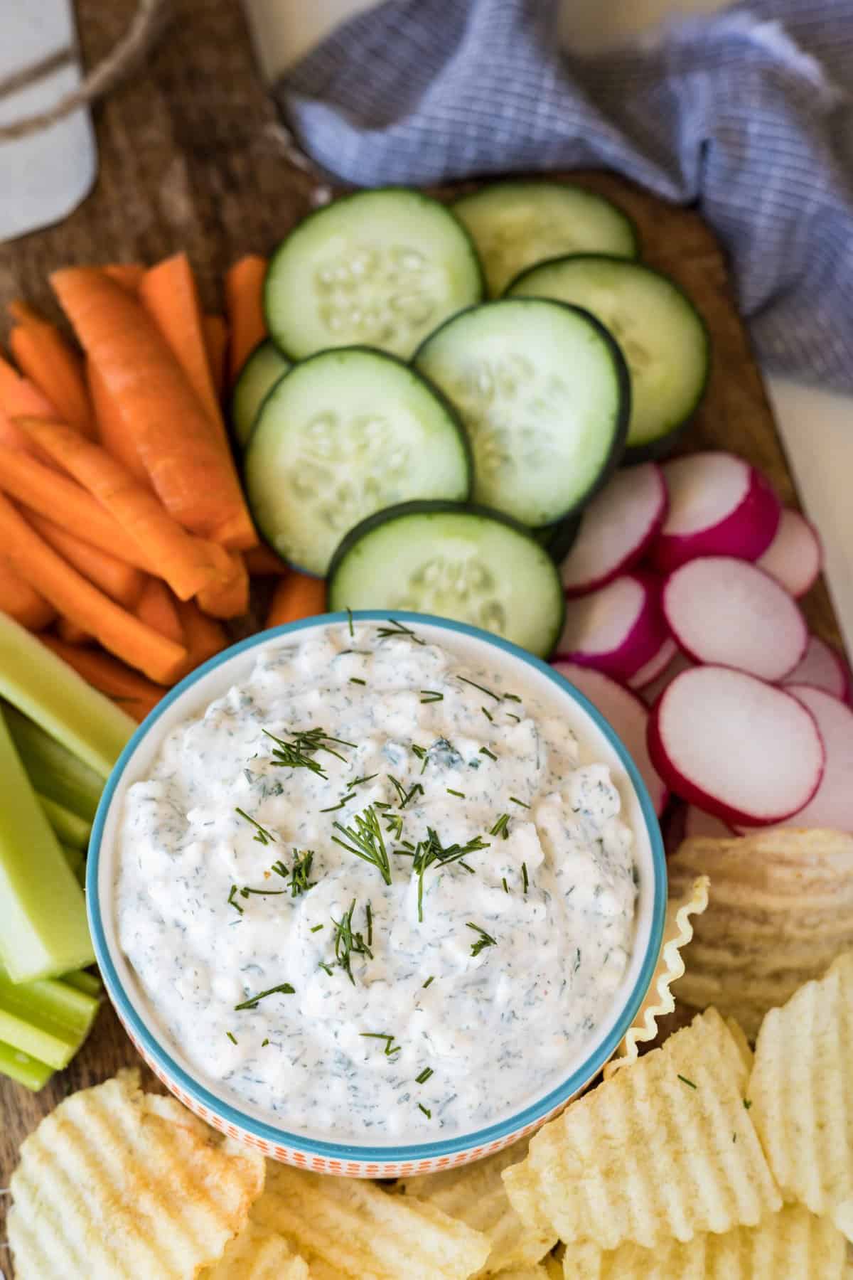 Fresh veggies and potato chips on a wooden board with veggie dip.