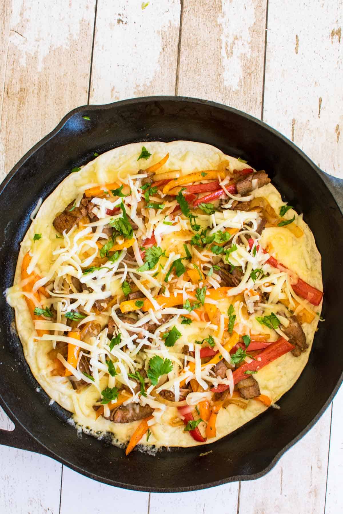 A tortilla in a skillet with sliced peppers, onions, and steak, sprinkled with grated cheese.