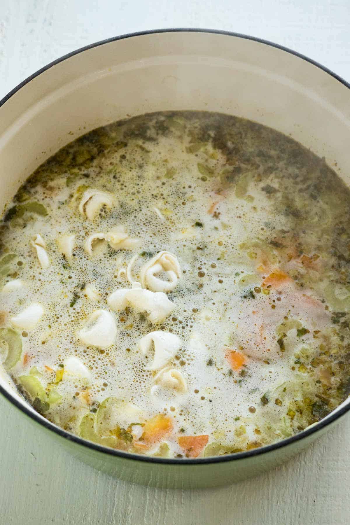 A large pot with boiling chicken broth and frozen tortellini noodles and carrots.