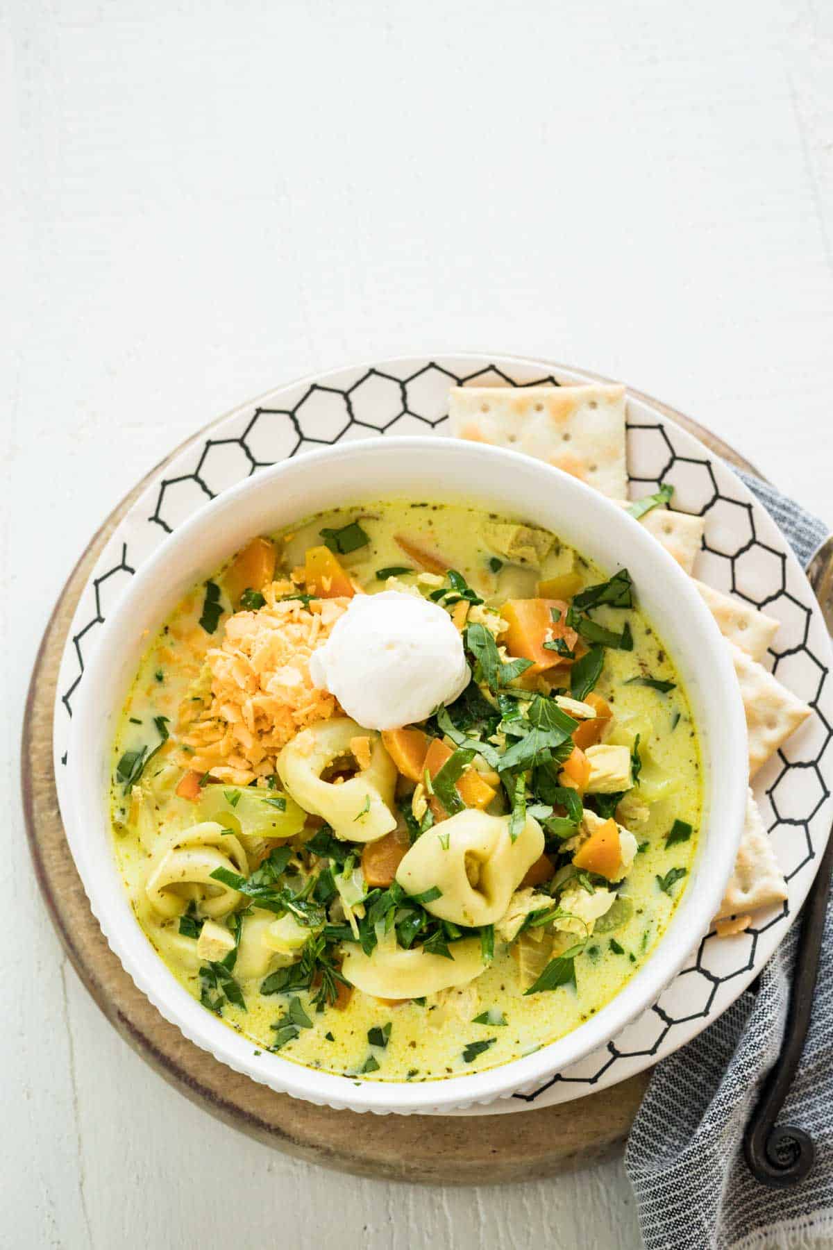 A bowl of soup with tortellini, spinach, and carrots topped with grated cheddar cheese and sour cream.
