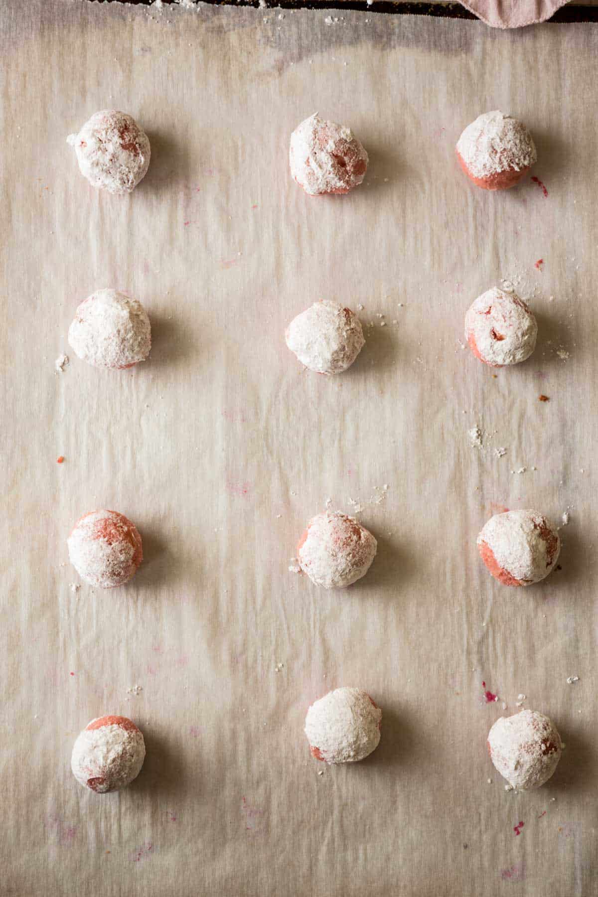 Balls of strawberry cookie dough rolled in powdered sugar on a parchment lined baking sheet.