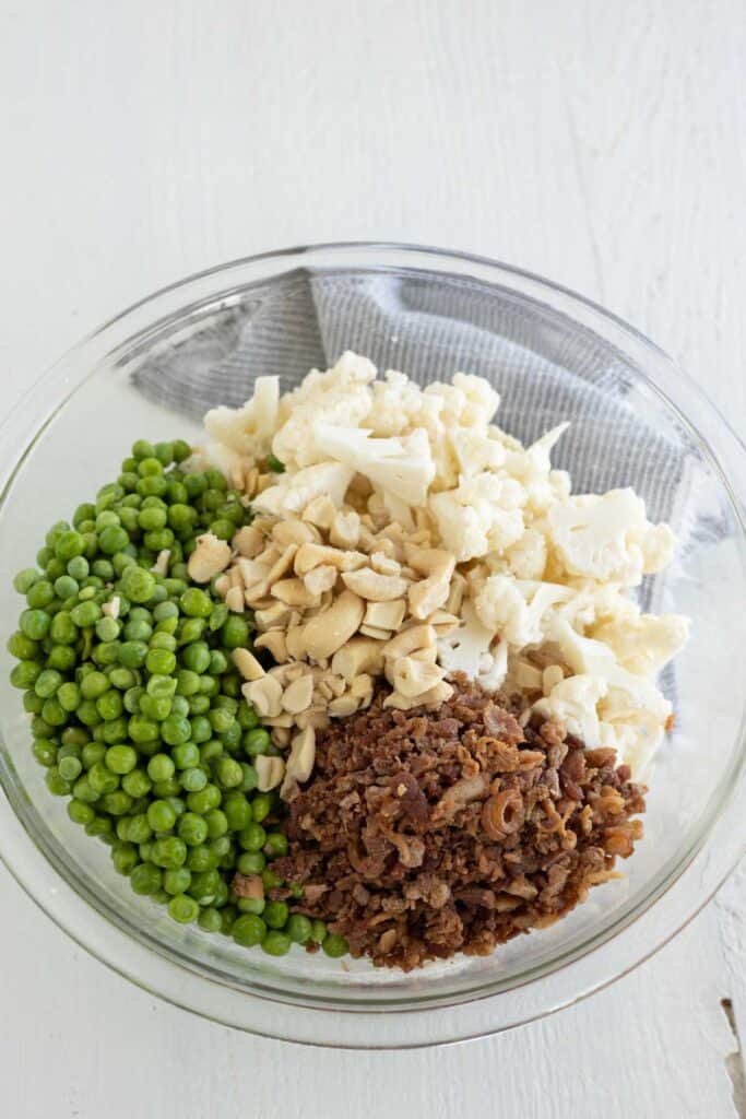 A large mixing bowl with bite size cauliflower florets, crumbled bacon, chopped cashews, and green peas.