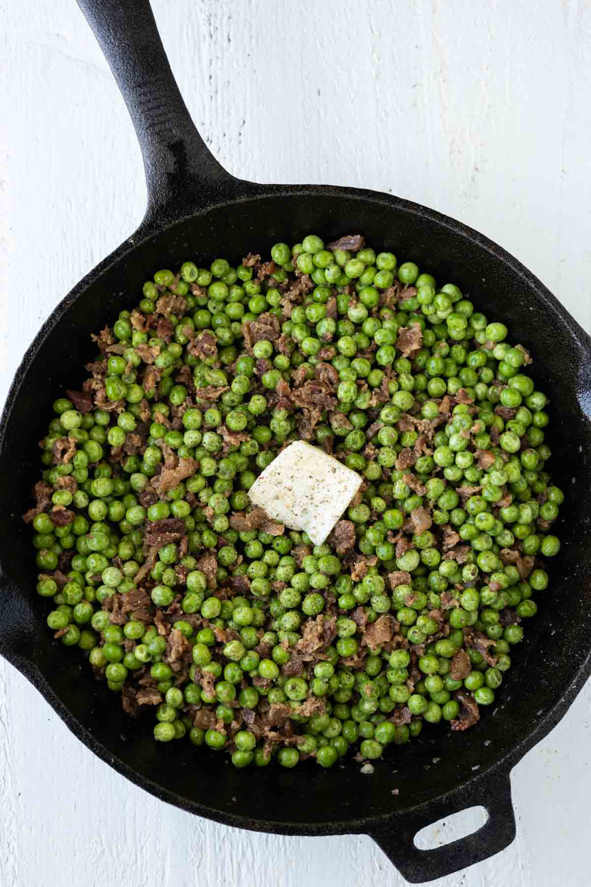 A skillet with cooked peas with bacon pieces and a pat of butter on top.