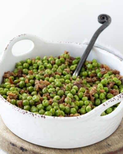 Green peas with bacon pieces in a white serving dish with a spoon.