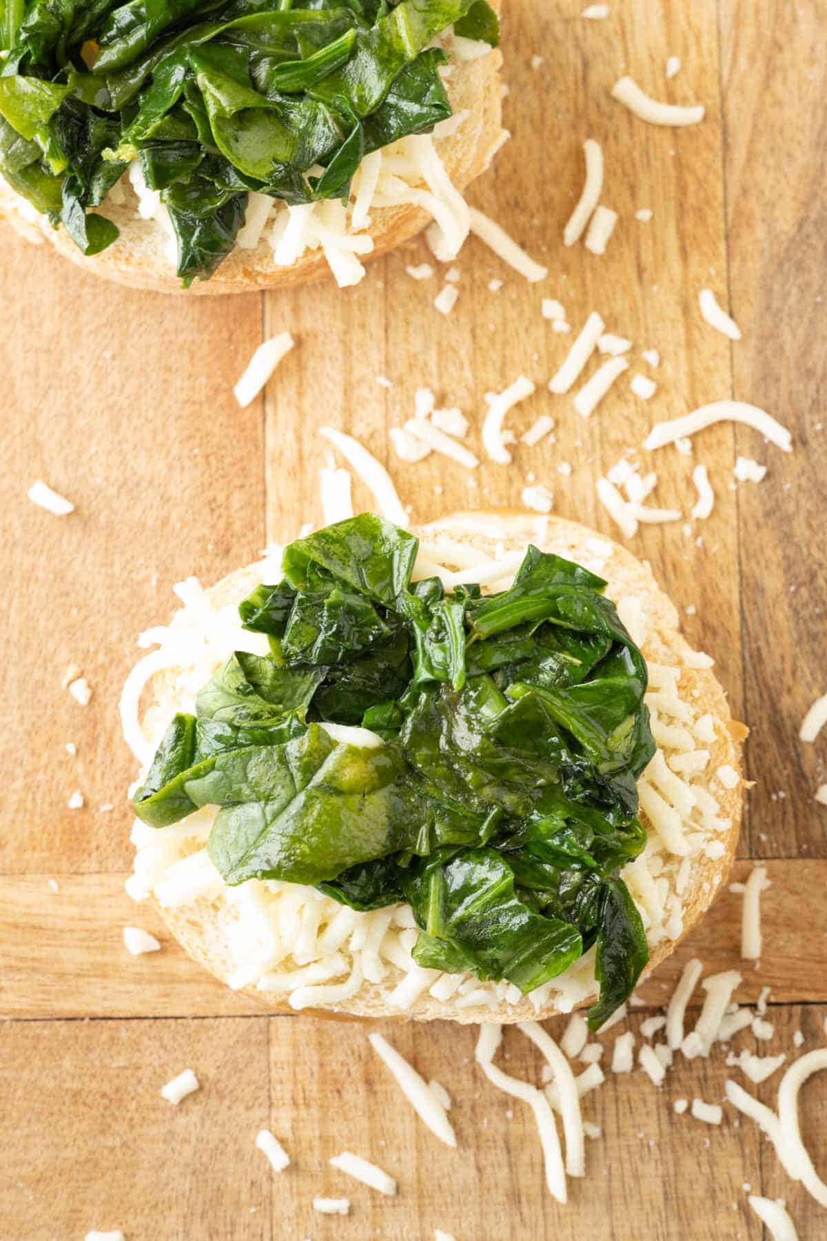 Slices of bread topped with grated cheese and sautéed spinach on a wooden cutting board.