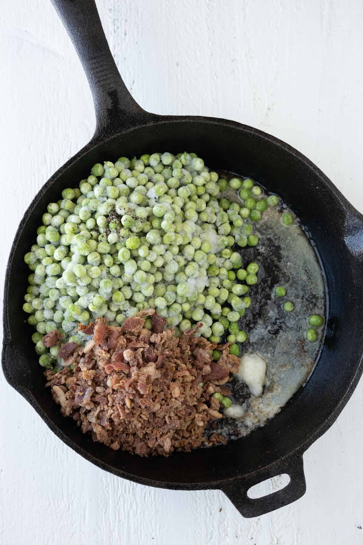 Frozen green peas, fried bacon pieces, butter, and garlic paste in a skillet.