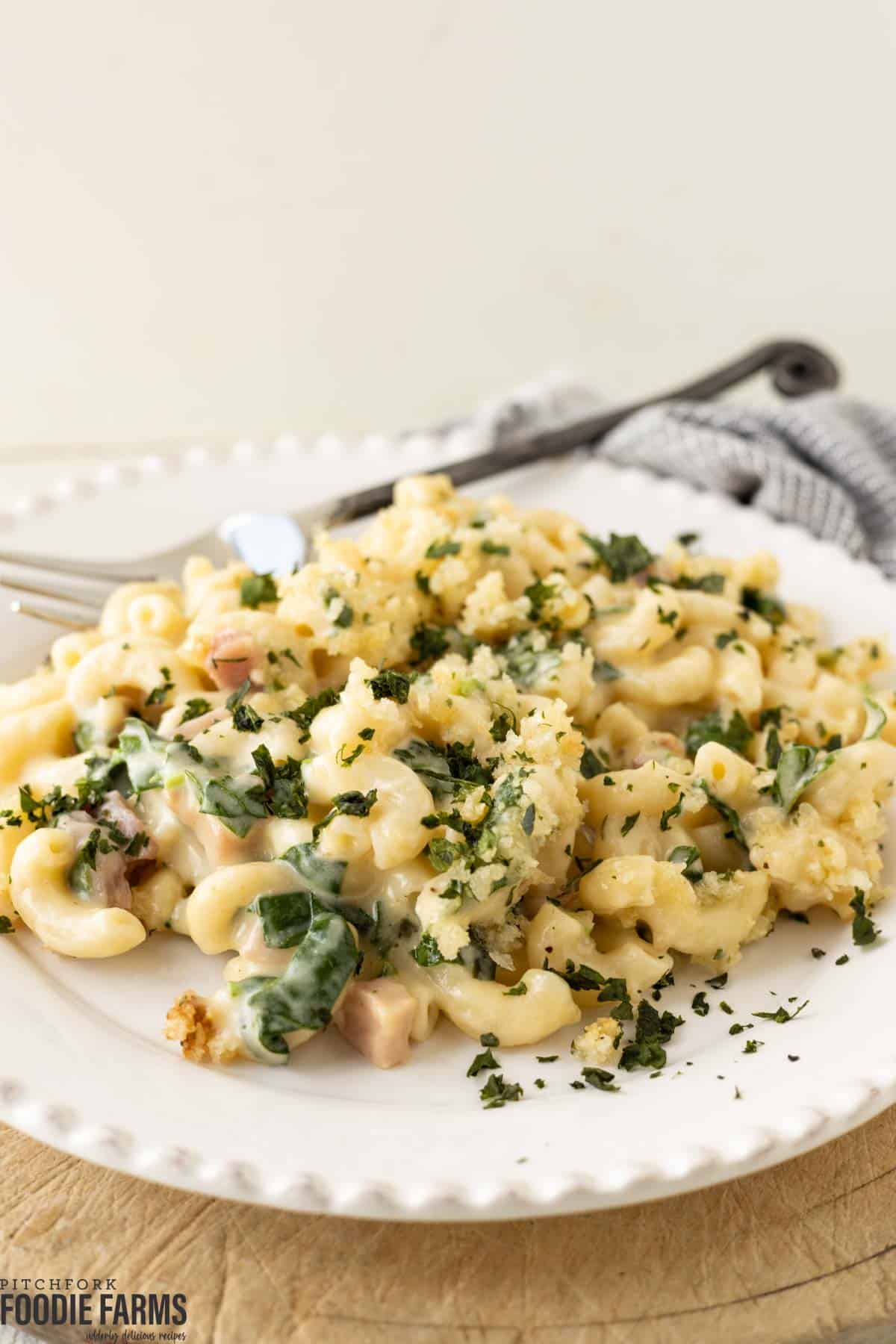 A white plate with homemade mac and cheese with chopped green spinach, diced ham, and a bread crumb topping.
