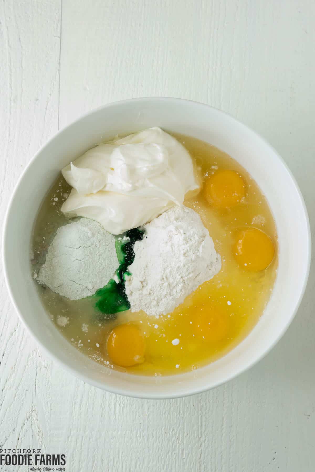 Cake mix, sour cream, eggs, food coloring, and pudding mix in a a white mixing bowl.