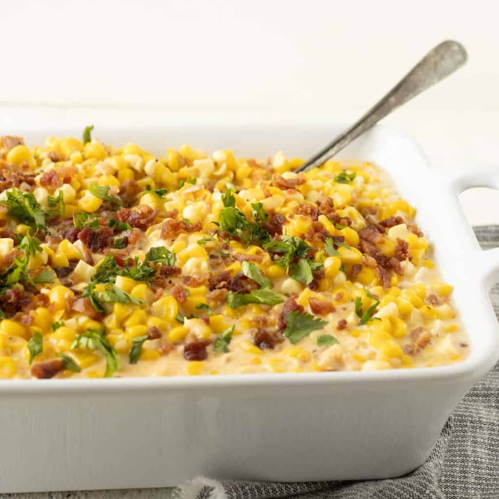 A dish of cheesy and cream corn with bacon and parsley on top.