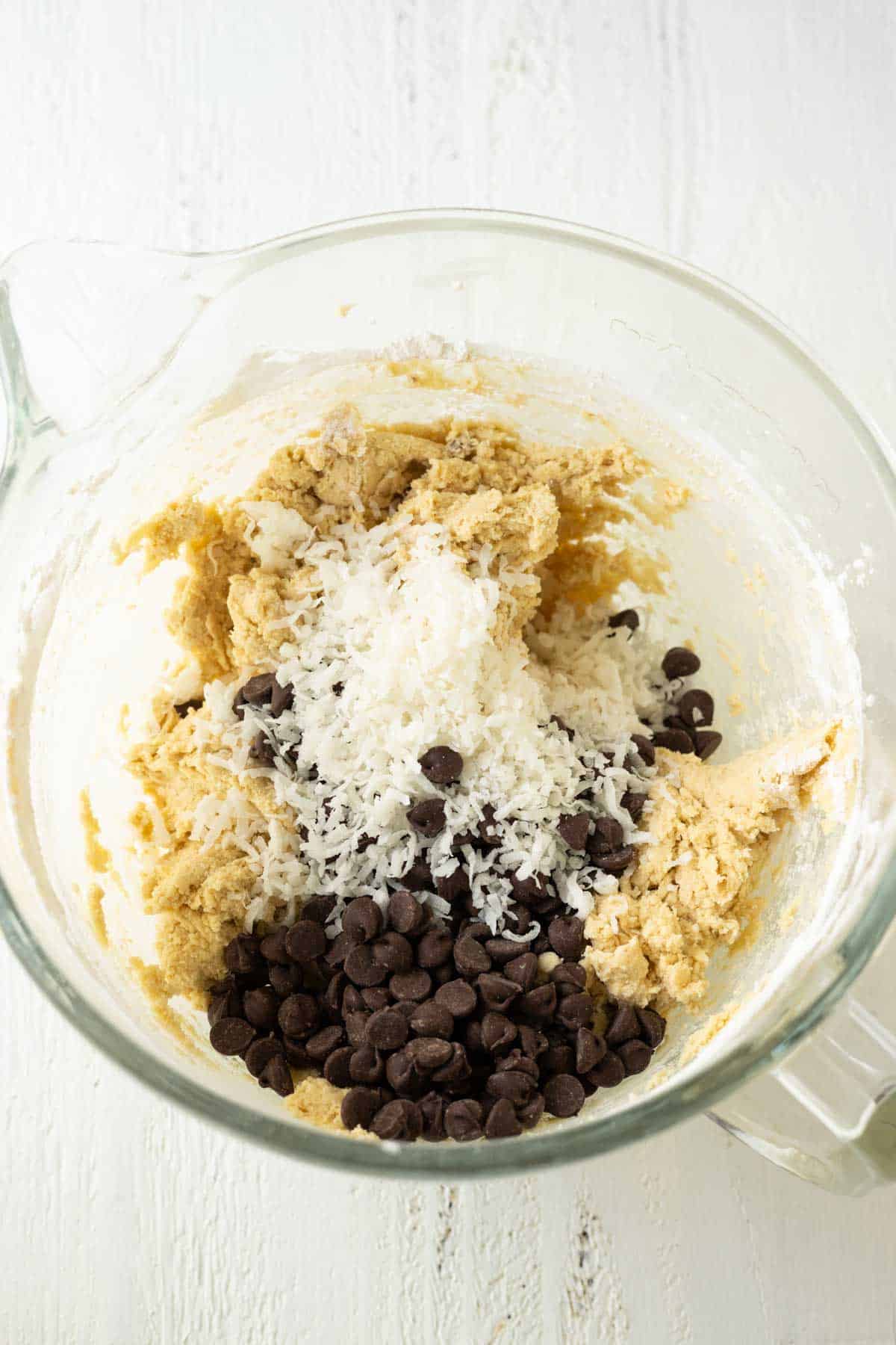 A mixing bowl with cookie dough and shredded coconut and semi sweet chocolate chips sprinkled on top.