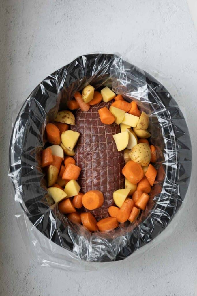 A plastic lined CrockPot with a roast beef, diced carrots, and potatoes.