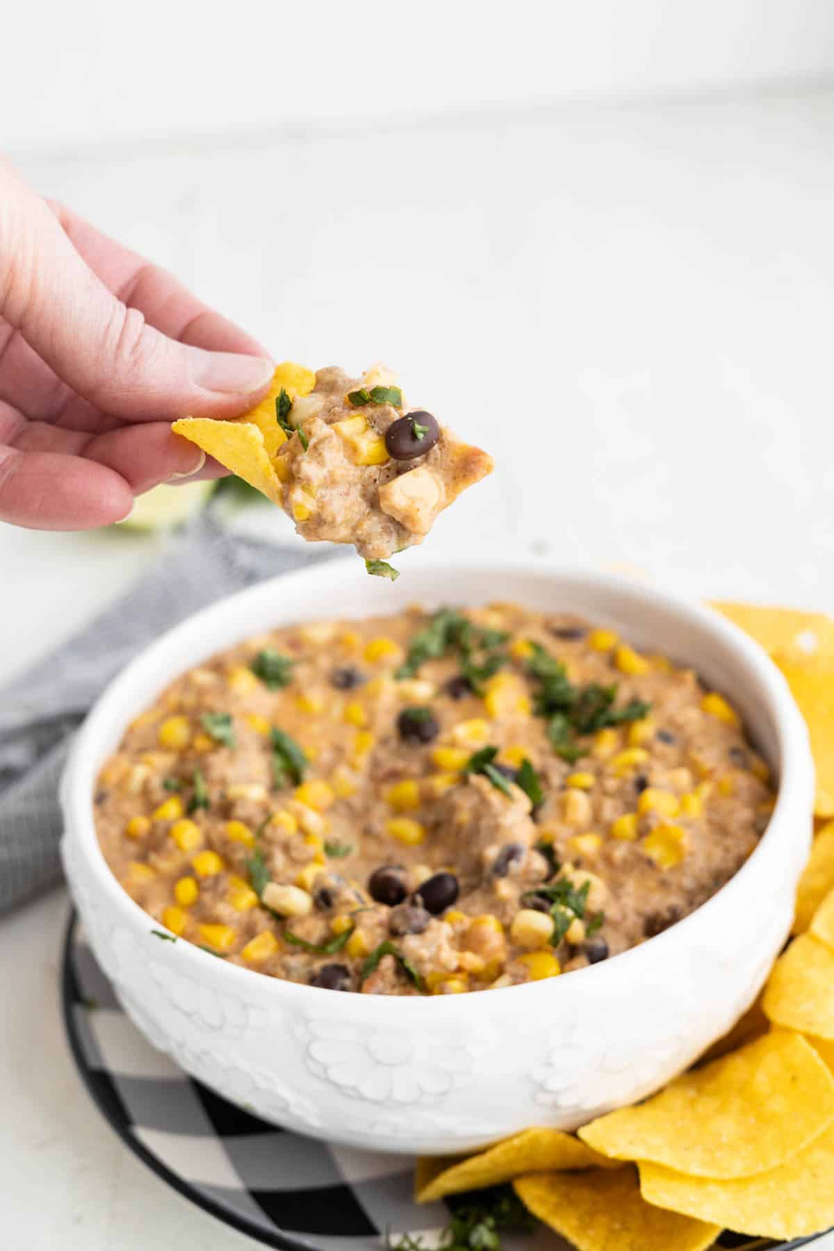 A bowl of creamy taco dip with corn, beans, and ground beef with a tortilla chip getting a bite.