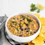 Taco dip with corn, ground beef, and beans in a white serving bowl with tortilla chips around it.