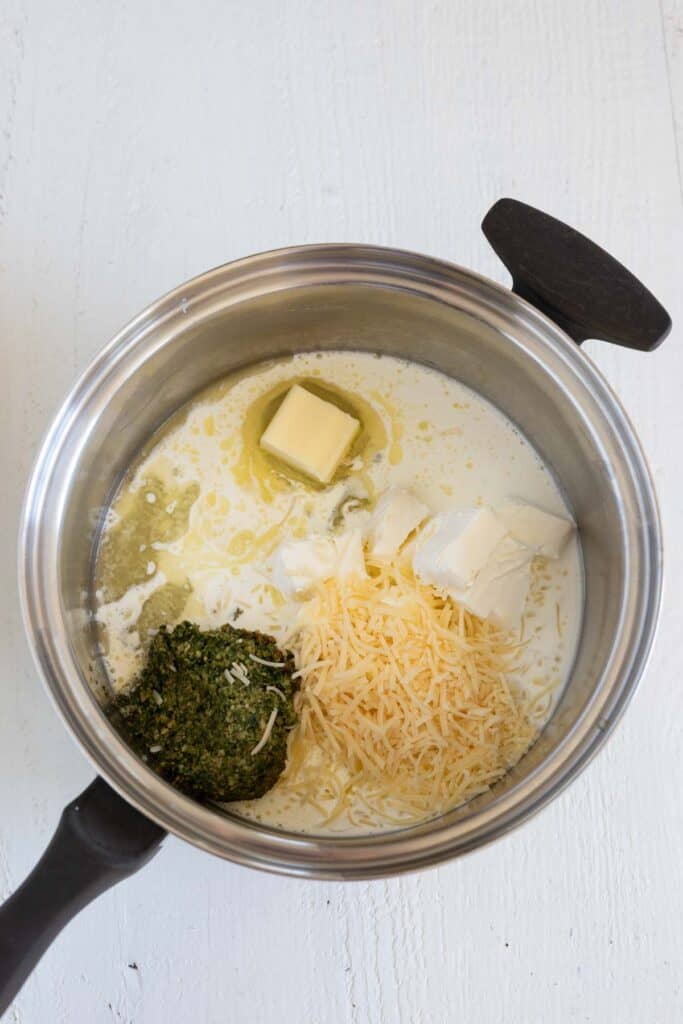A saucepan with butter, cream, grated parmesan cheese, cream cheese, and pesto in it. 